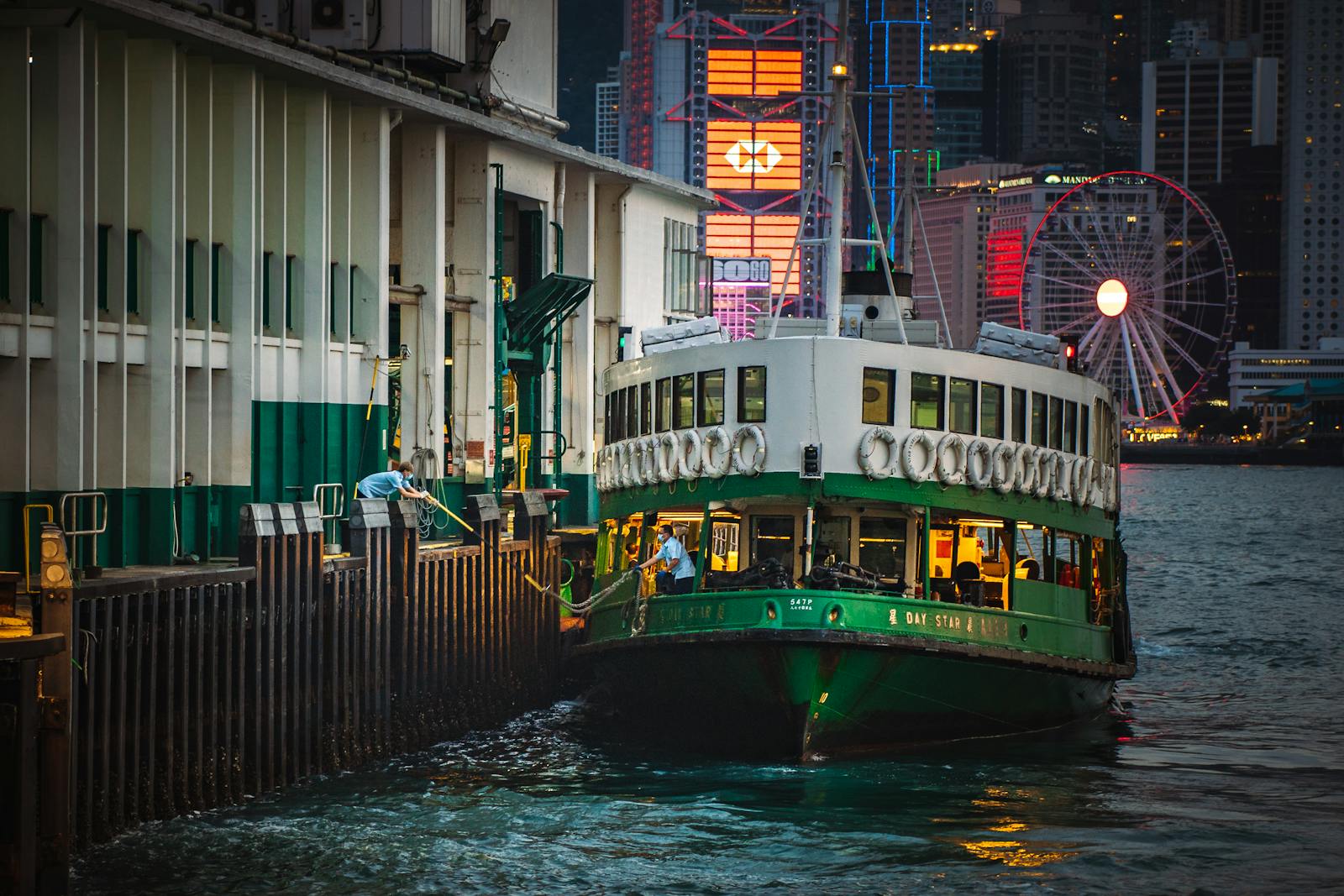 The Star Ferry boat docked at night with Hong Kong skyline and ferris wheel in the background.