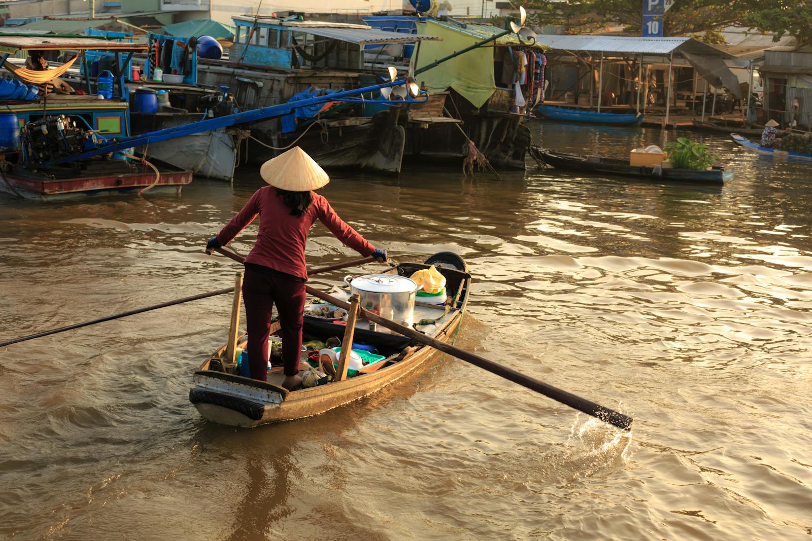 Asian woman rows a boat at a bustling river market, showcasing traditional water transport.