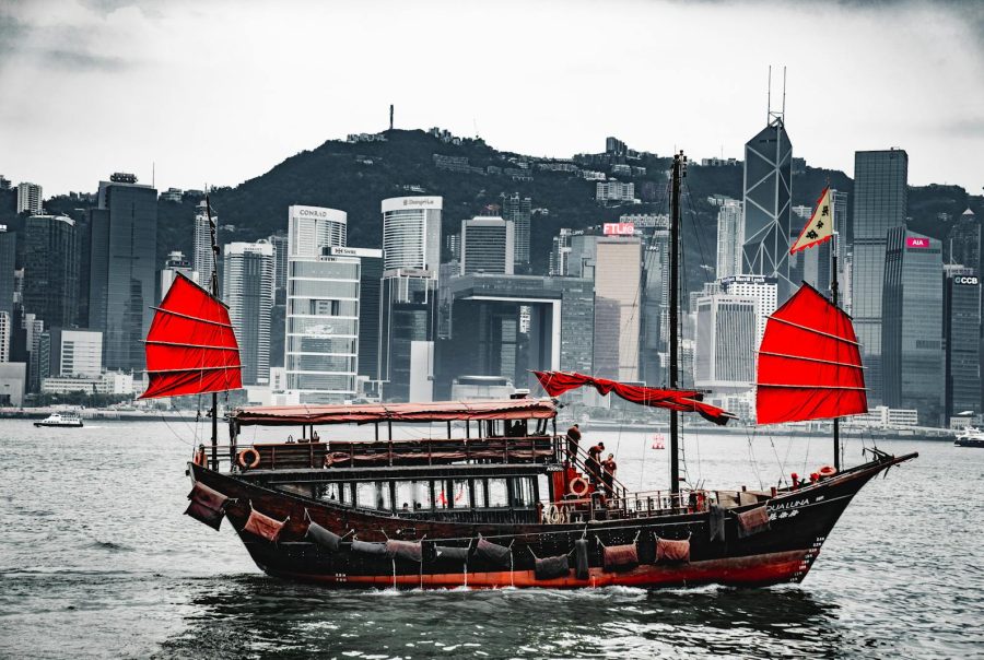 A traditional junk boat with red sails in Hong Kong harbor, against a skyline backdrop.