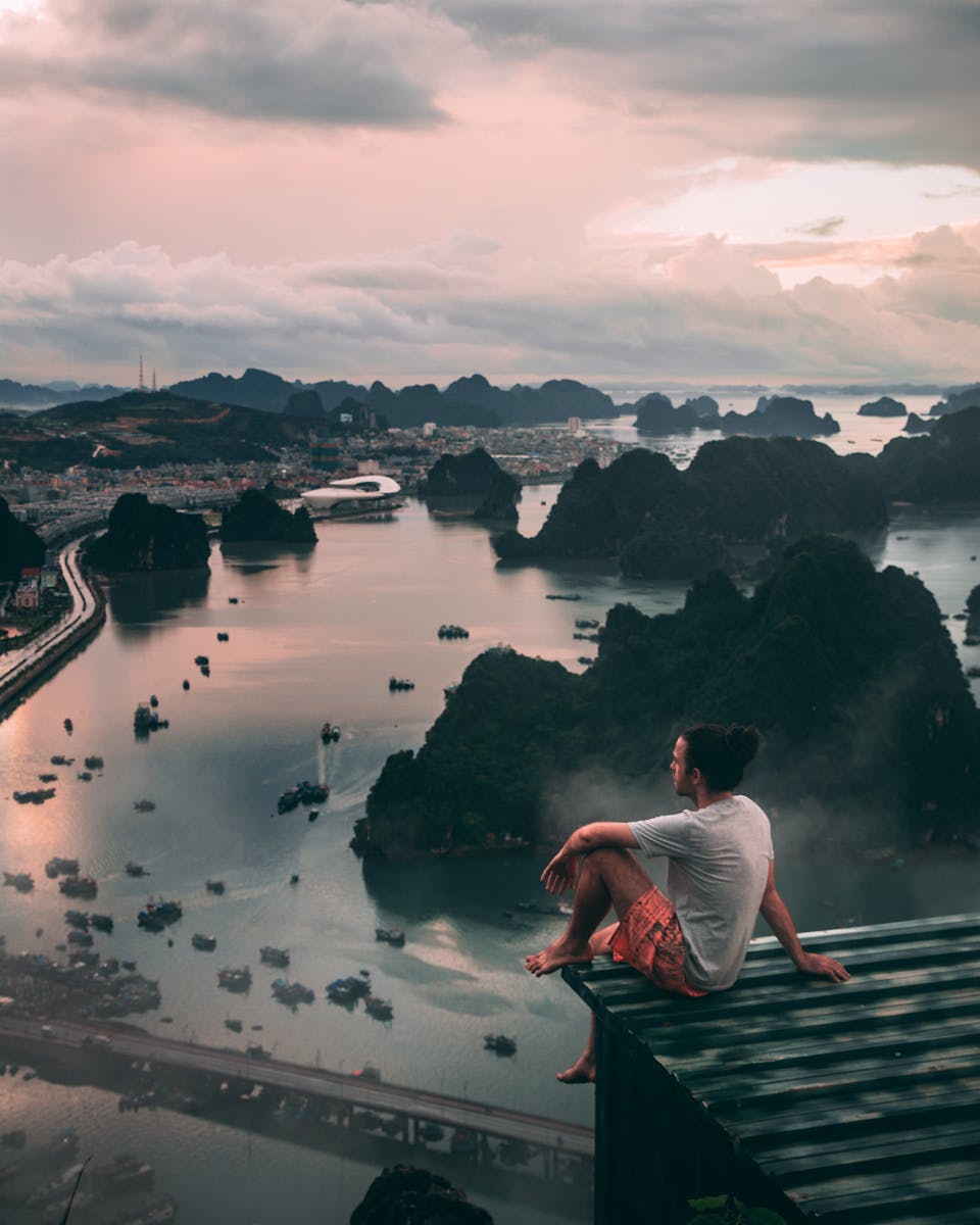 Man sitting on rooftop overlooking Halong Bay's stunning landscape during sunset.
